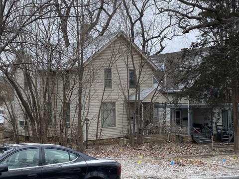 A black car is parked in front of a house with bare trees.