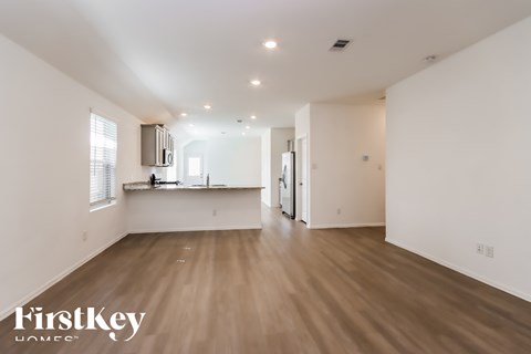 A spacious room with wooden flooring and white walls, with a kitchen area visible in the background.