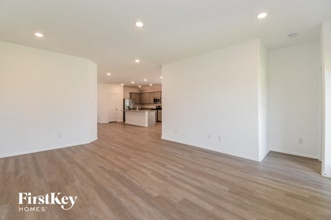 A spacious room with wooden flooring and white walls, with a kitchen visible in the background.