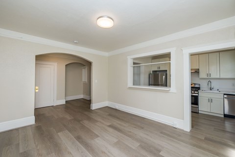 A kitchen with a refrigerator, oven, and sink is visible through a window in a room with wooden flooring.