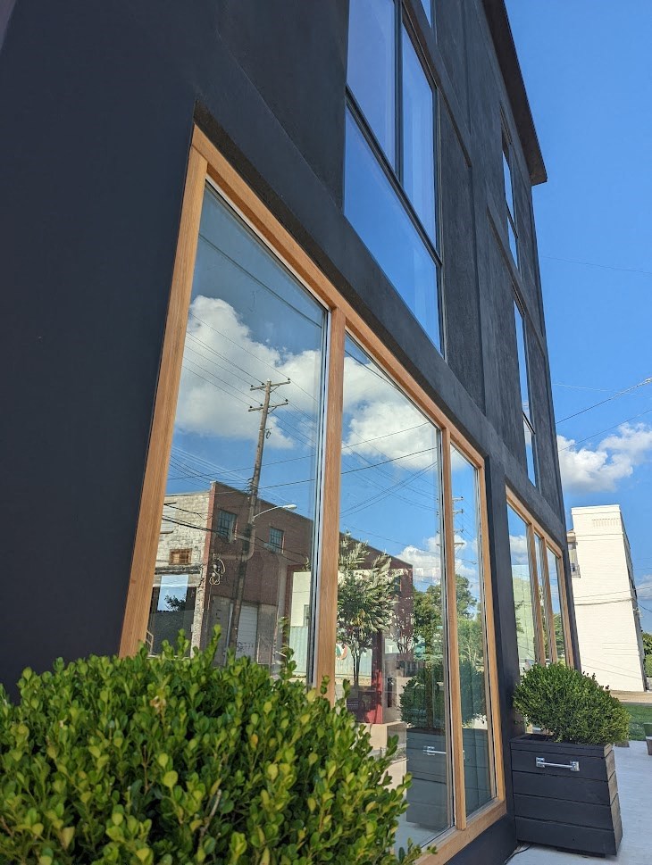 A building with a dark grey exterior and a large window reflecting the sky and a building.