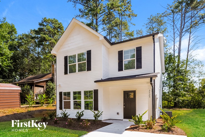 A white house with black shutters and a black door is surrounded by greenery.