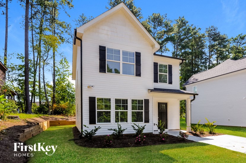 A white house with black shutters and a black roof is surrounded by greenery.