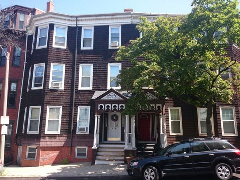 A black car is parked in front of a brown building with a red door.