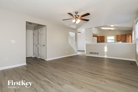 A spacious room with a ceiling fan and light fixture, wooden flooring, and a staircase leading to another room.