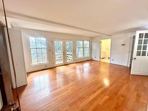 A room with wooden floors and white walls, featuring a large window with a view of trees and a door leading to another room.