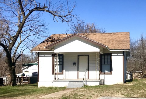 A small white house with a brown roof and a tree in front.