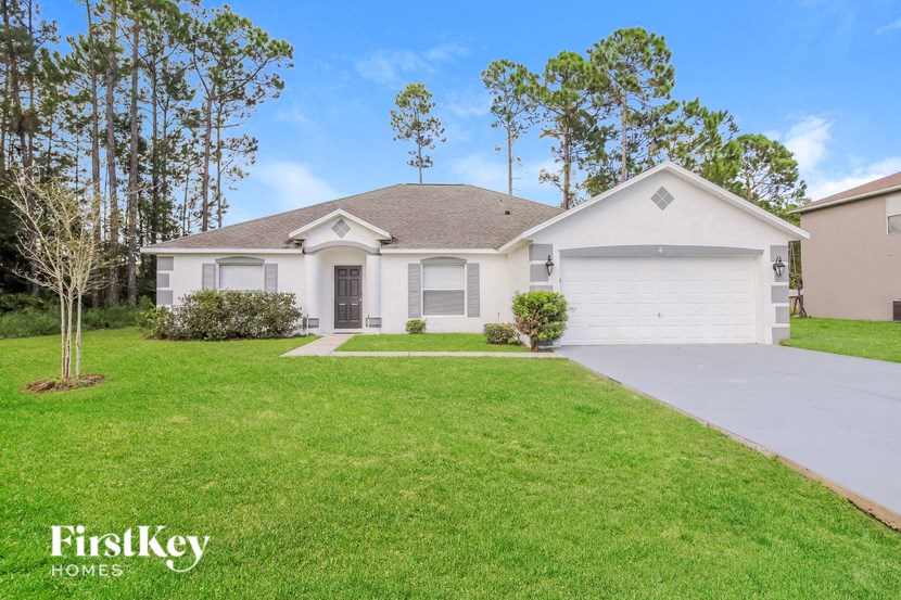 A white house with a brown roof and a garage door is surrounded by a grassy lawn and trees.