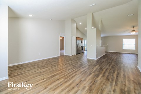 A spacious room with wooden flooring and white walls, with a kitchen visible in the background.