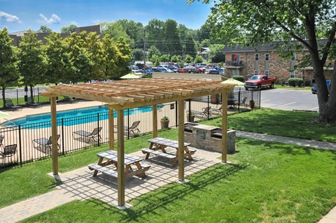 A wooden picnic table is in the foreground of a grassy area with a pool and a covered picnic area in the background.
