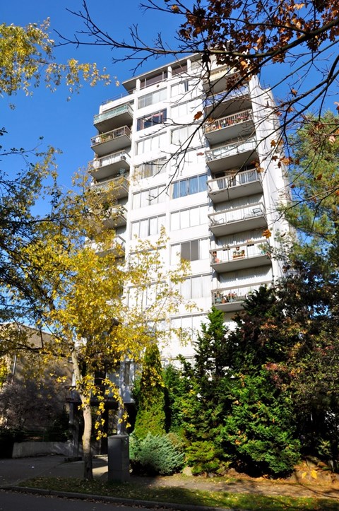 A tall white building with balconies is surrounded by trees.