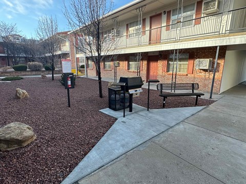 A courtyard with a bench, a trash can, and a tree.