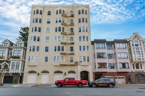 A red truck is parked in front of a beige building with a black car behind it.