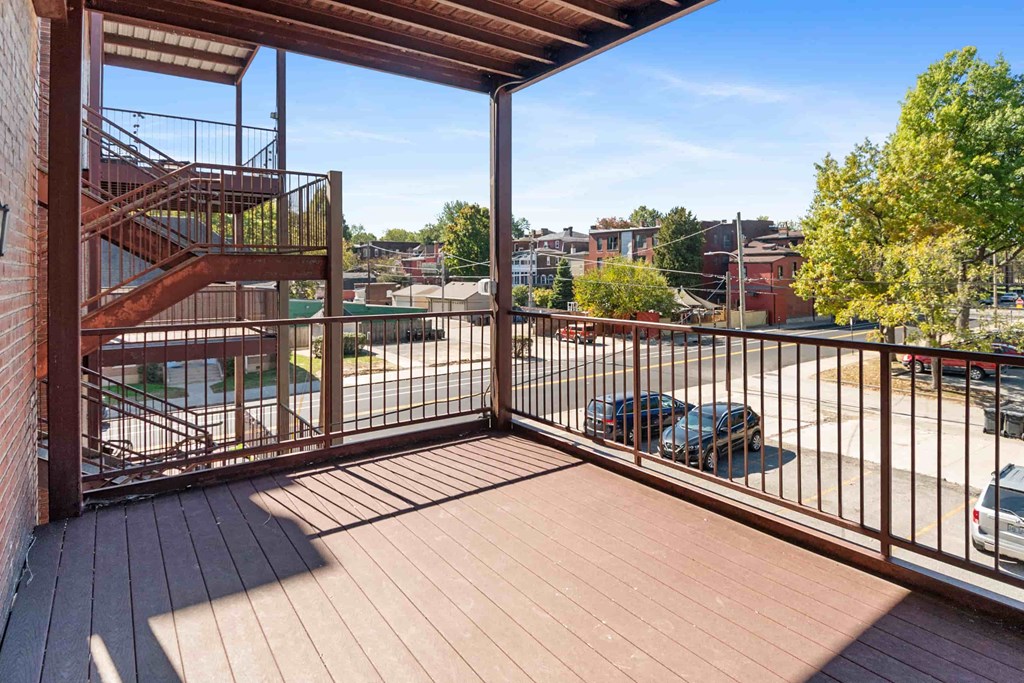 A balcony with a metal railing and a wooden floor overlooks a parking lot and buildings.