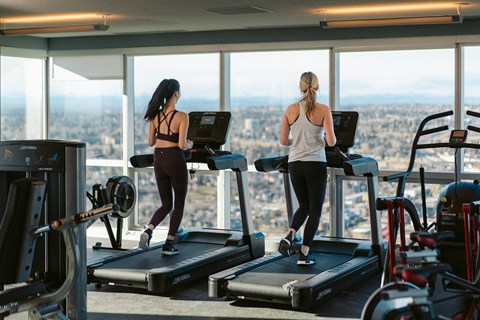 two women running on treadmills in a gym overlooking the city