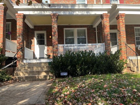 a front porch of a brick house with a white railing and a yard with bushes