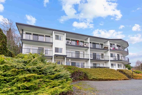A white building with balconies and windows is surrounded by greenery.