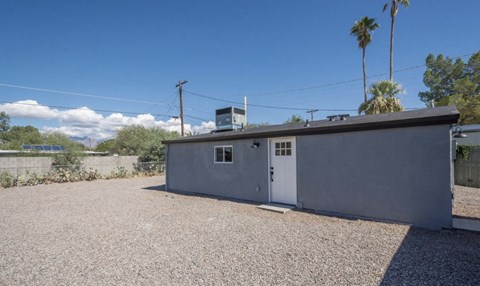 A small grey building with a white door and a window is surrounded by a gravel lot.