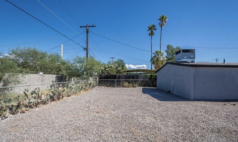 A gravel driveway leads to a house with a fence and palm trees in the background.