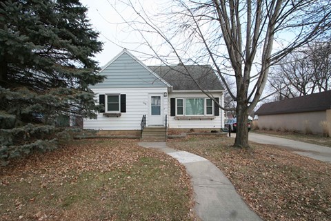 A small house with a grey roof and white siding is surrounded by trees and a sidewalk.