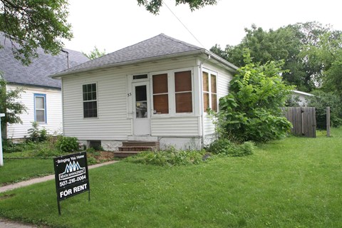 A small white house with a black sign in front that says "For Rent".