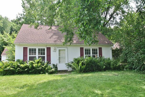 A small white house with red trim and a red door numbered 368 is surrounded by greenery.