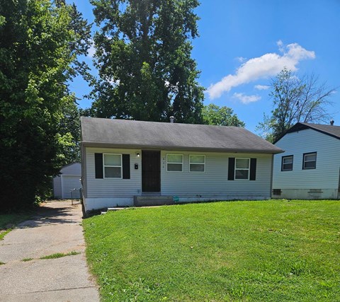 A small house with a grey roof and white siding is surrounded by a green lawn and trees.