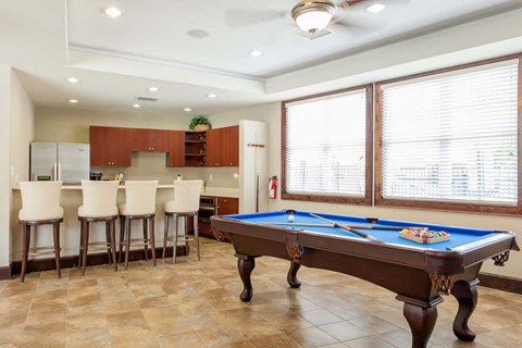 A pool table is in the middle of a room with a bar and a kitchen in the background.