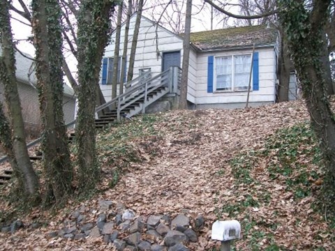 A small house with a blue and white exterior is surrounded by trees and a stone wall.