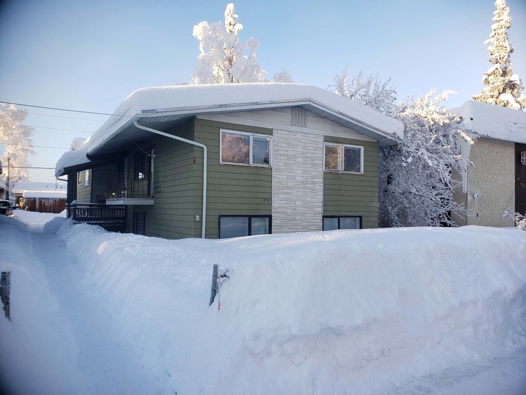 A house with a green siding and a grey roof is surrounded by a large amount of snow.