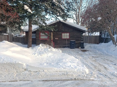 A wooden cabin with a red wreath on the window is surrounded by snow.