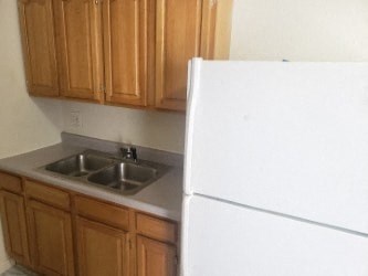 A white refrigerator stands next to a kitchen sink with wooden cabinets above it.
