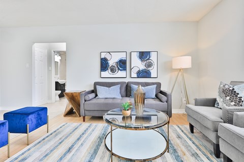 A living room with a grey couch, a blue ottoman, a glass coffee table and a painting on the wall.
