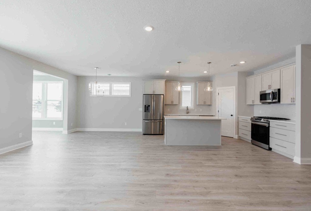 an empty living room with a kitchen with a refrigerator and a sink