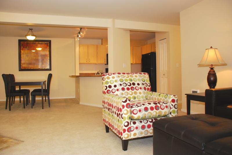 A living room with a black chair, a black ottoman, a black table, a black refrigerator, a black cabinet, a black lamp, a painting, a dining table, and a dining table with chairs.