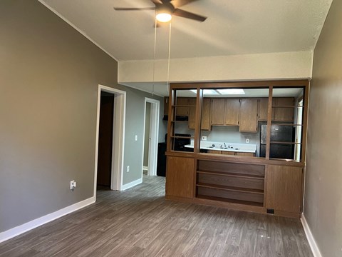 A room with a ceiling fan and a kitchen area in the background.
