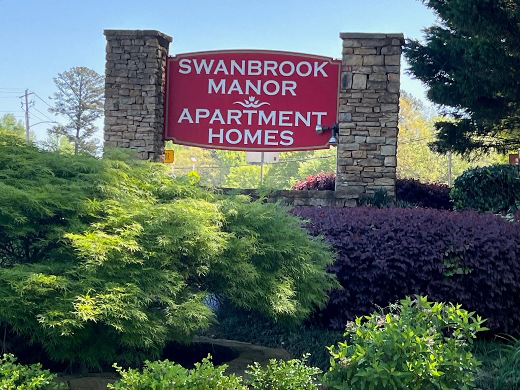A sign for Swanbrook Manor Apartment Homes is displayed in front of a lush green landscape.