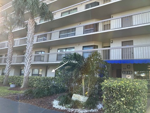 A building with balconies and a blue awning is surrounded by greenery.