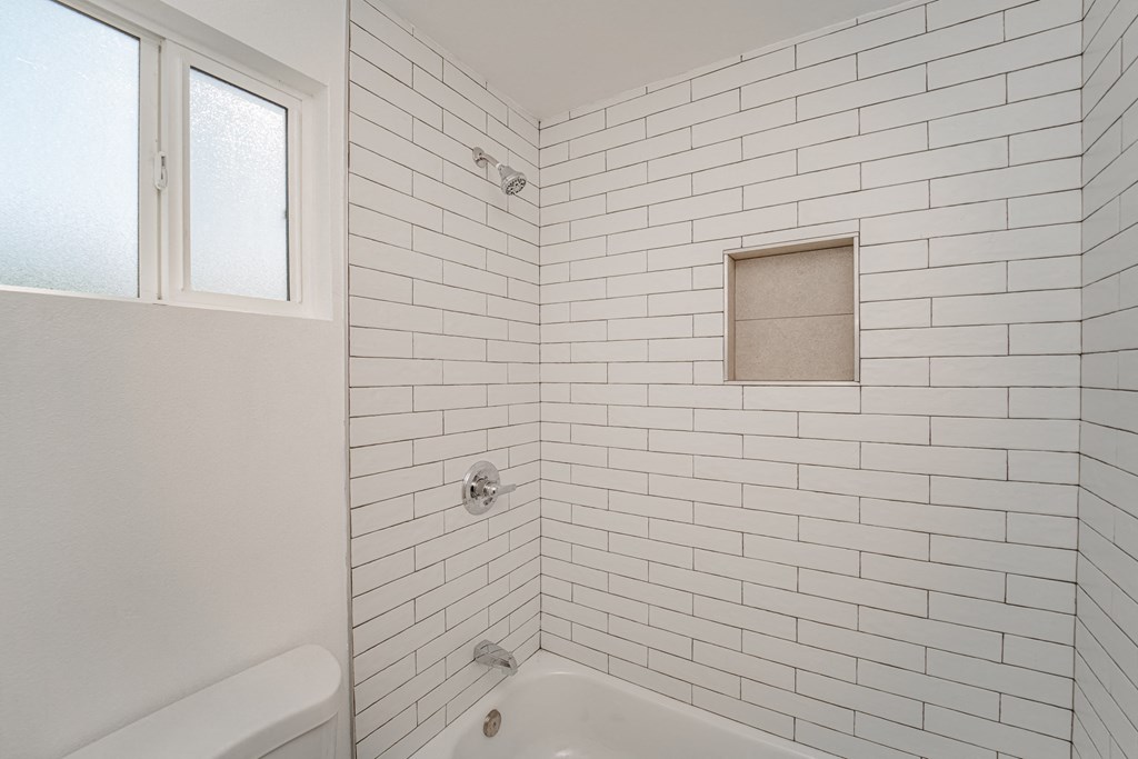 a white tiled bathroom with a tub and shower and a window