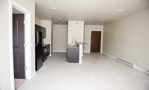 an empty living room with a kitchen and a black refrigerator
