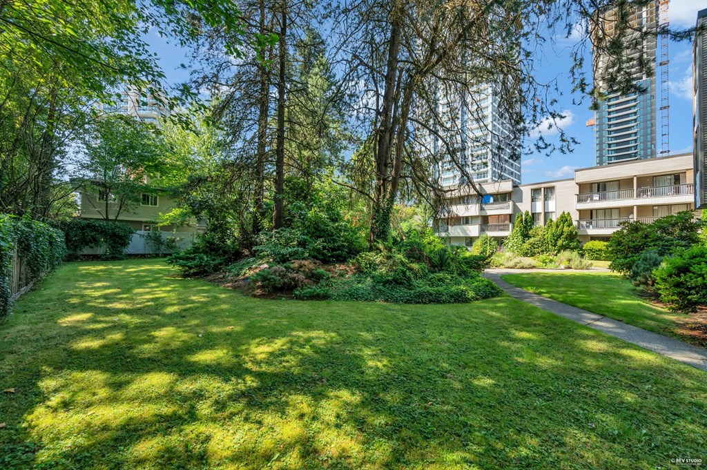 A lush green garden with a pathway and trees in the foreground and apartment buildings in the background.