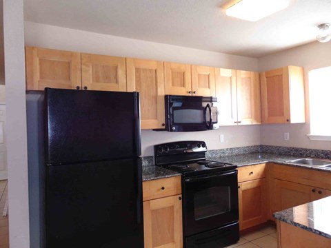 A black refrigerator stands in a kitchen with wooden cabinets and granite countertops.