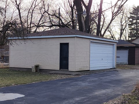 A small garage with a black door is surrounded by a driveway and trees.