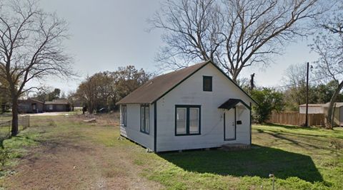 A small white house with a black roof and a black door is surrounded by a grassy area and trees.