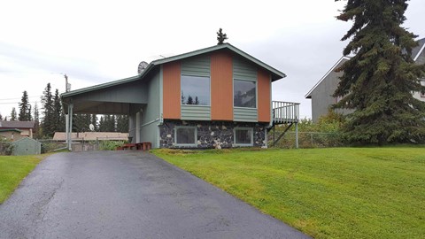 A house with a green and orange exterior sits on a grassy area with a driveway leading to the front door.