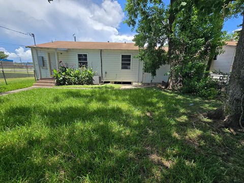 A small house with a brown roof and a white exterior is surrounded by a grassy area and trees.