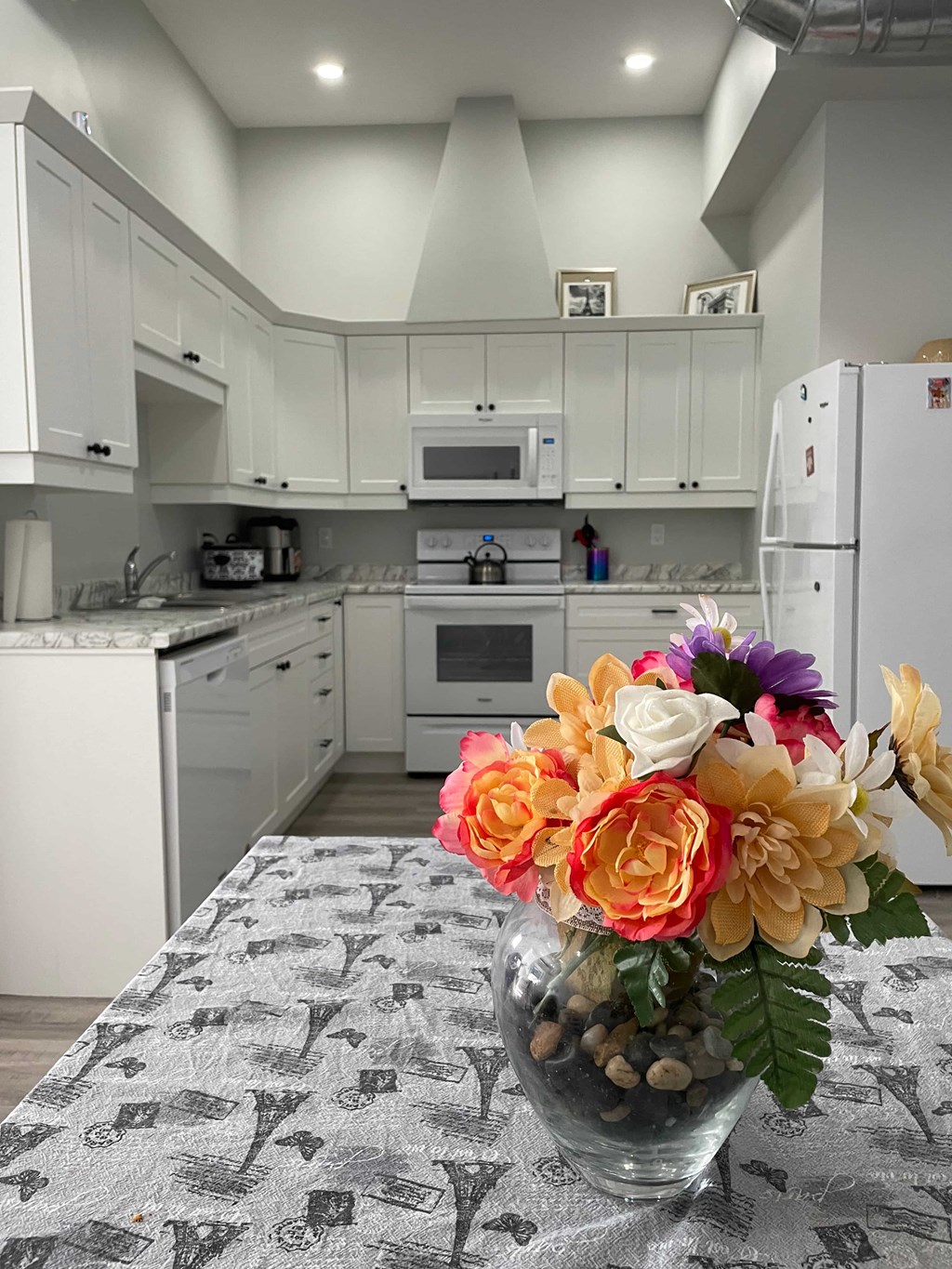 A kitchen with a table covered in a cloth with letters on it and a vase of flowers on the table.