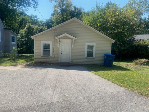 a small tan house with a driveway and a blue trash can