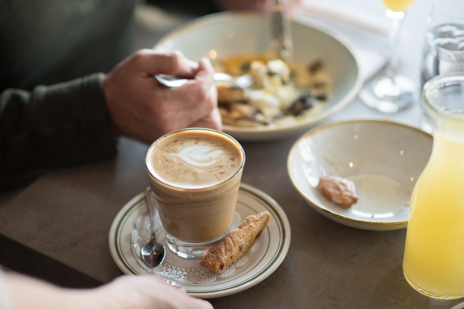 A person eating at a table with a latte, biscotti, breakfast food, and a cup of orange juice at a restaurant at Eagleview Town Center.