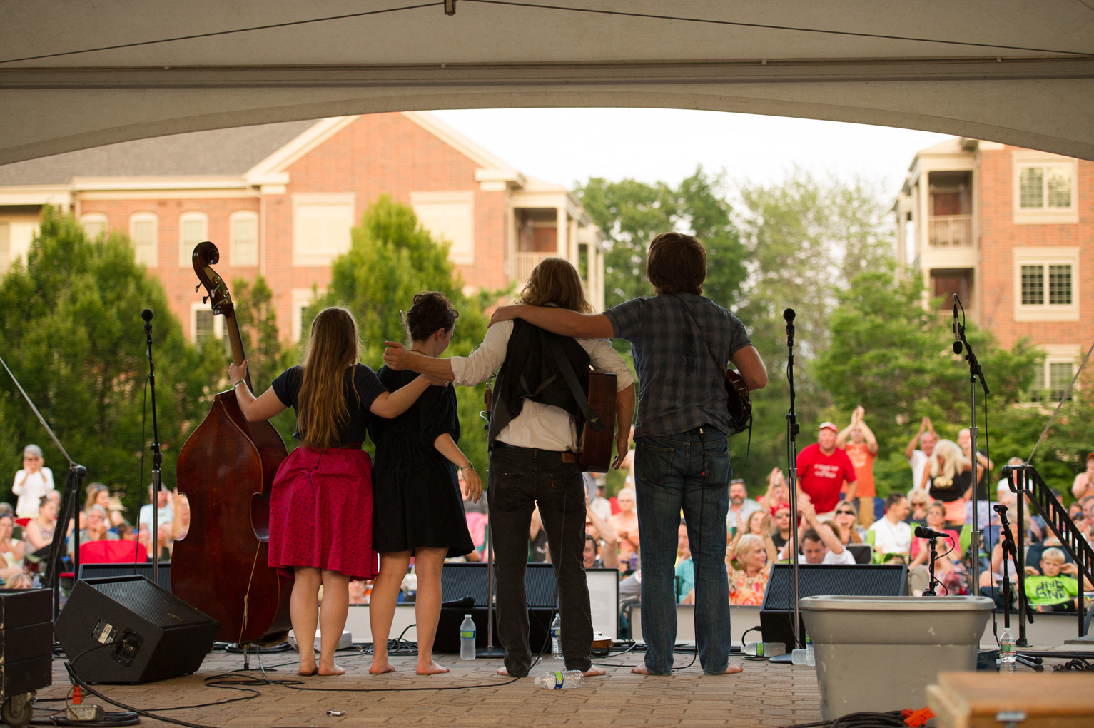 A concert at Eagleview Town Center showing the backside of four performers with their instruments on an outdoor covered stage with a crowd of people watching them.